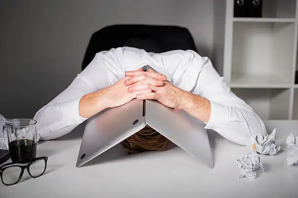 person slumped over desk with head under open laptop