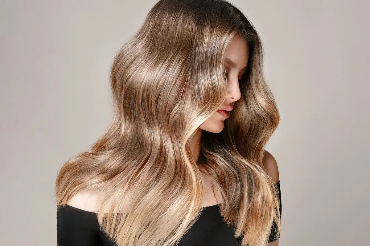 A woman with long, wavy, and healthy-looking brown-to-blonde balayage hair wearing a black off-the-shoulder top. A woman with long, wavy, and healthy-looking brown-to-blonde balayage hair wearing a black off-the-shoulder top.